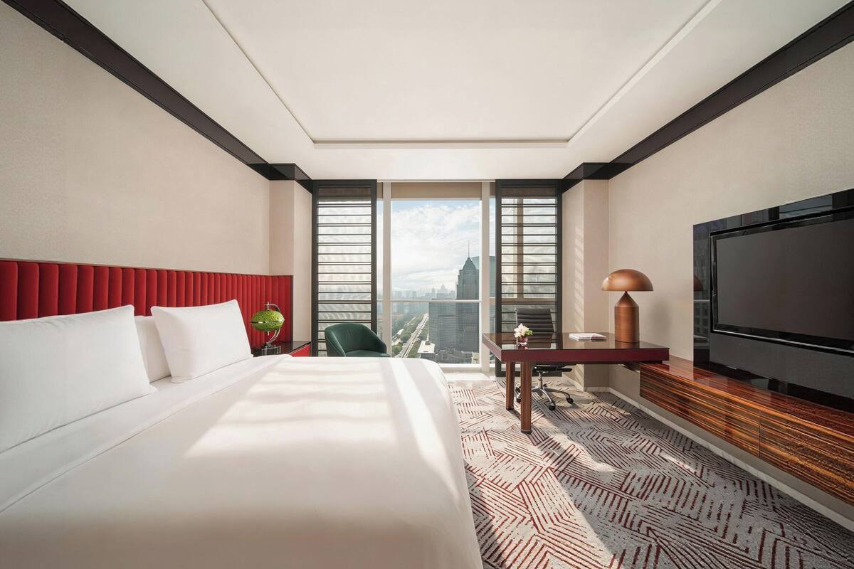 A hotel room with a large bed, a desk with a lamp, a TV, and a view of the cityscape.