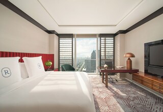 A hotel room with a large bed, a desk, a chair, a television, and a view of a cityscape through large windows.