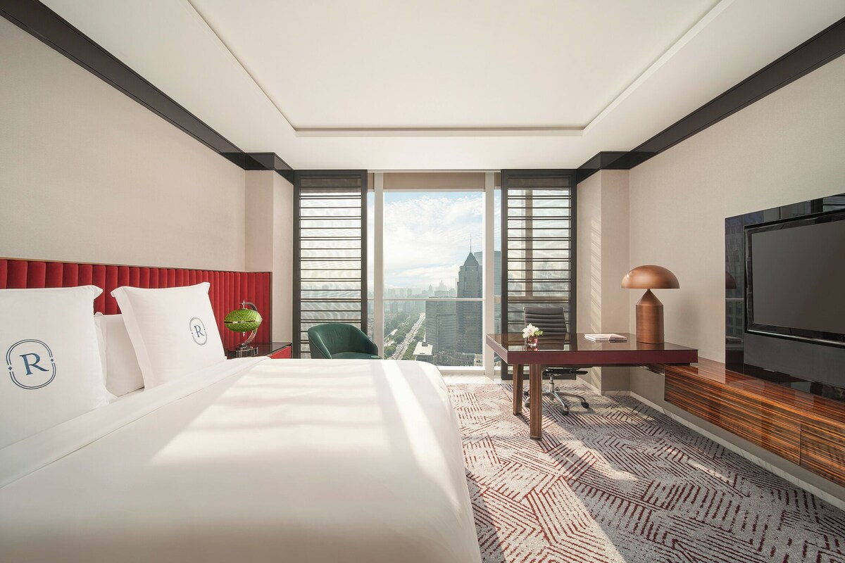 A hotel room with a large bed, a desk, a chair, a television, and a view of a cityscape through large windows.