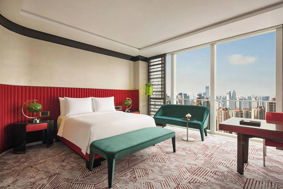 A hotel room with a large bed, a bench, a desk, a chair, and a view of a cityscape.