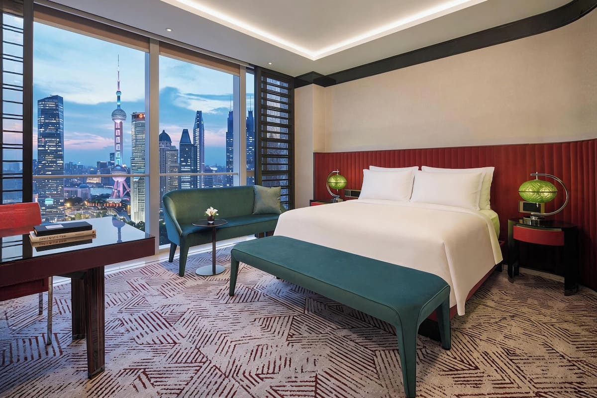 A hotel room with a large window, a bed, a green bench, a small table, and a nightstand with lamps.
