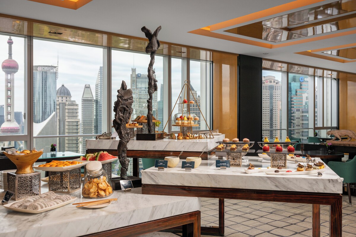 A buffet setup with various food items and a decorative tree sculpture in a modern hotel room with large windows overlooking a cityscape.