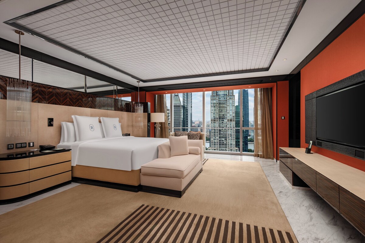 A modern hotel room with a large bed, a sofa, a TV, and a city view.