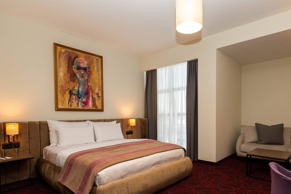 A hotel room with a bed, a sofa, a chair, a painting, and a window with curtains.