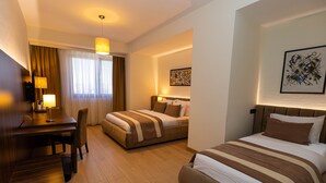 Una habitación de hotel con escritorio, dos camas, zona de comedor y un ventanal con cortinas.