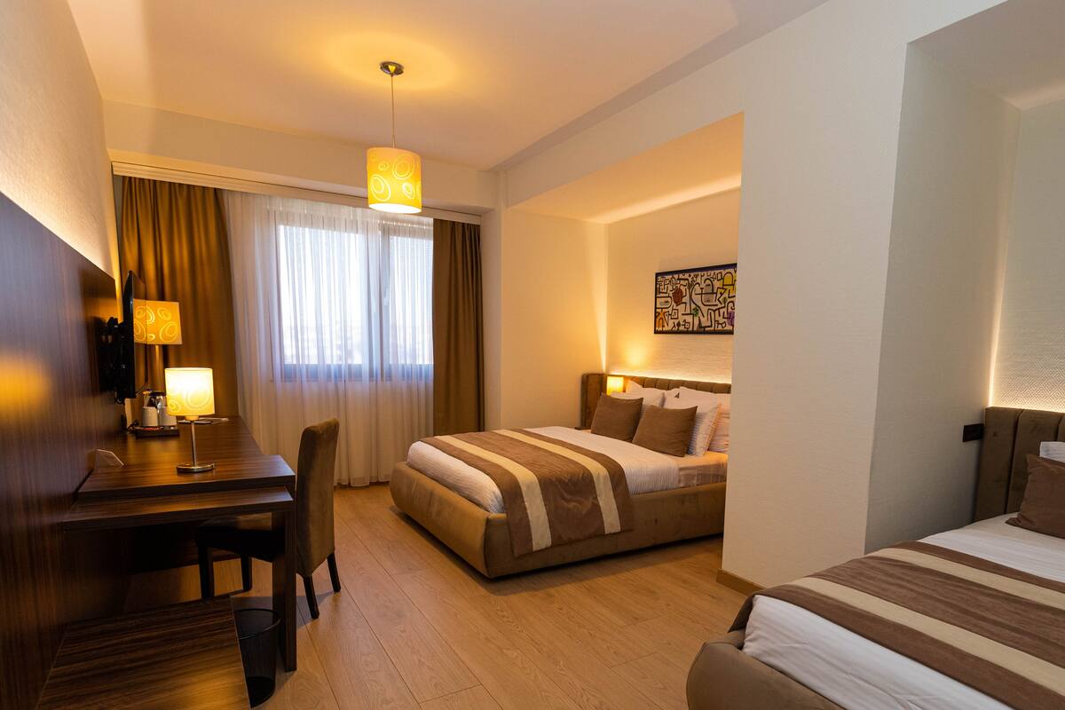 A hotel room with a desk, two beds, a dining area, and a large window with curtains.