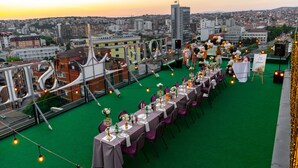 Montaje para un evento al aire libre con mesas y sillas en una azotea, con vistas a un paisaje urbano al atardecer.