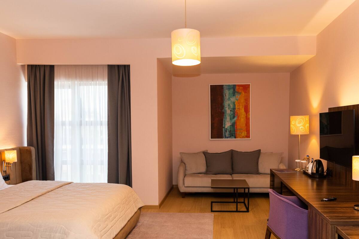 A hotel room with a bed, a sofa, a desk with a lamp, a TV, and a painting on the wall.