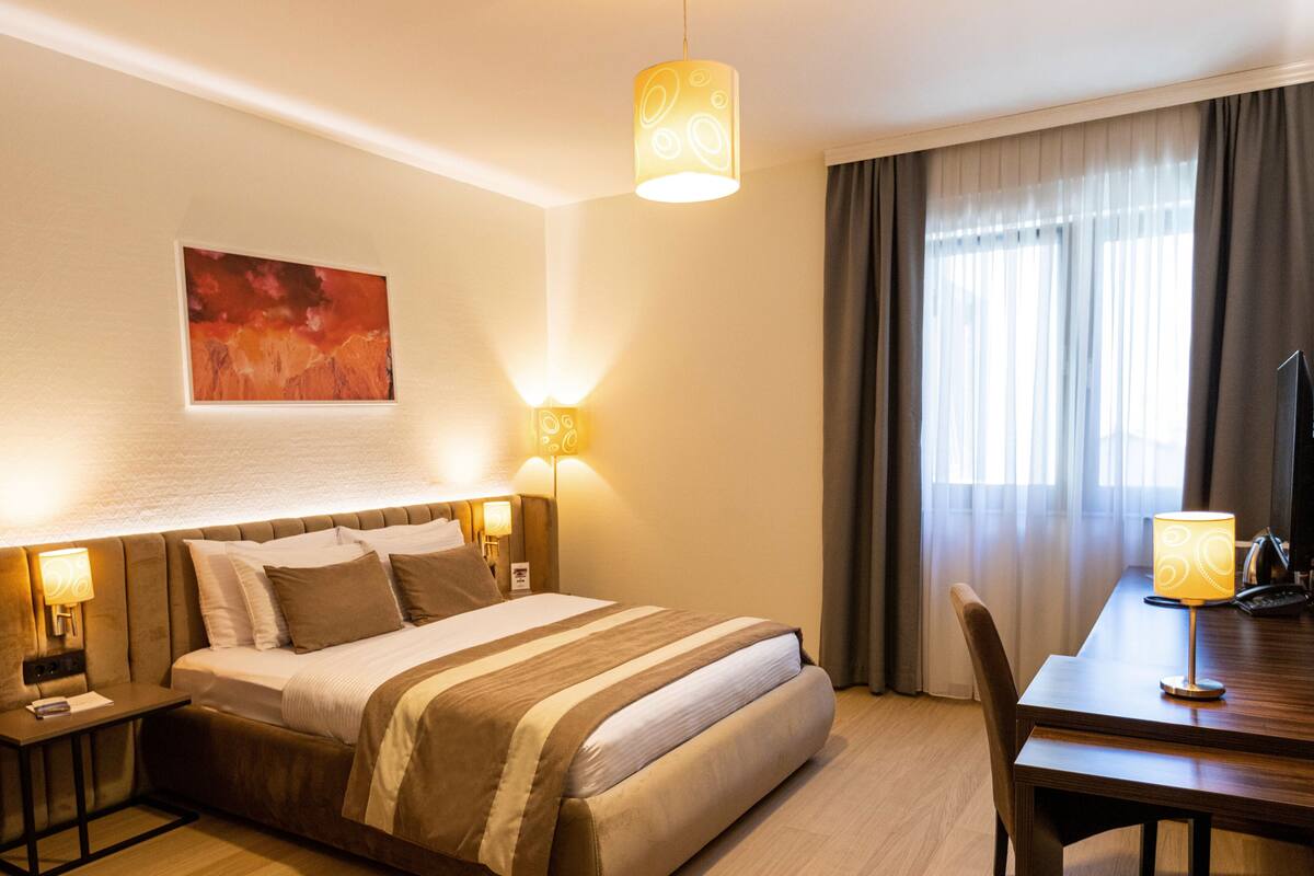 A hotel room with a bed, bedside tables, a desk with a chair, a television, and a large window with curtains.