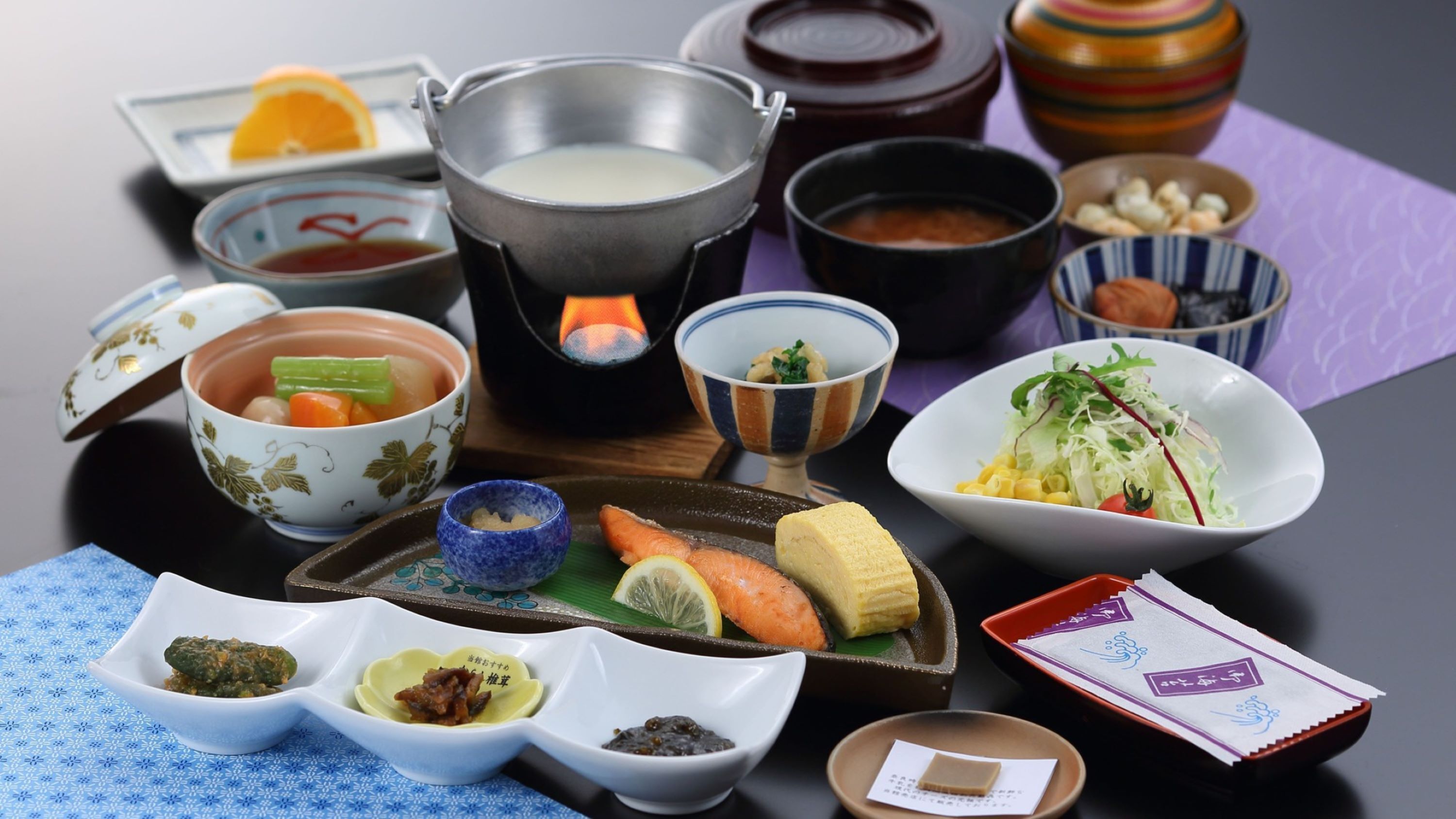 A variety of Japanese dishes including sushi, sashimi, and a pot of soup, arranged on a table with various small bowls and a teapot.