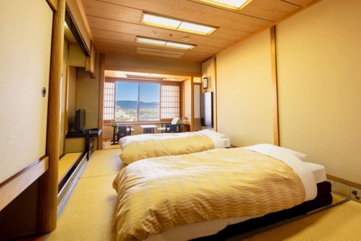 A hotel room with two beds, a desk, a chair, and a large window offering a view of the cityscape.