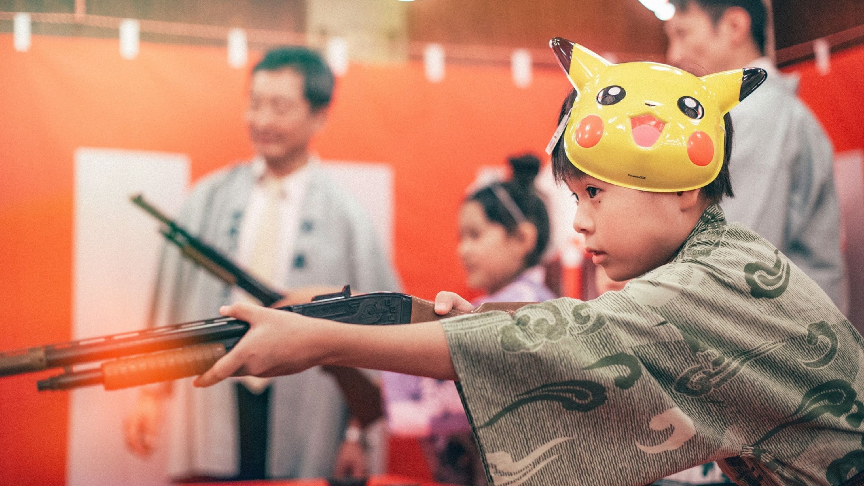 Una persona vestida con traje tradicional sostiene una pistola de juguete, con un gorro de Pikachu, en una habitación con paredes rojas y una puerta blanca.