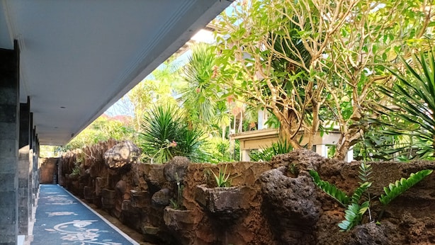 A covered walkway with a blue patterned floor, flanked by a stone wall and lush greenery. - Sapta Petala Hotel (Kuta)