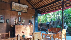 A wooden reception area with a counter, framed artwork, and a clock. There are wooden chairs and tables, and a stone wall with a statue in the background. - Sapta Petala Hotel (Kuta)