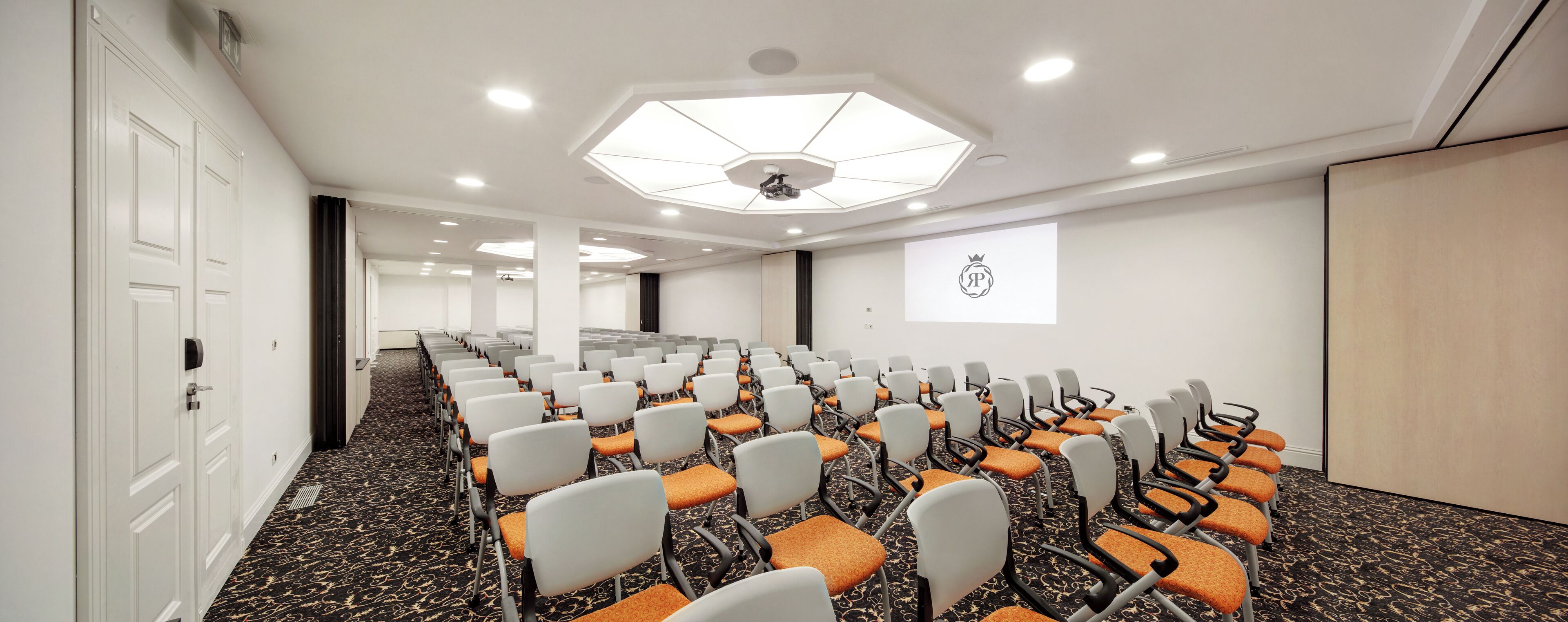 Konferenssihuone, jossa on riveissä tuoleja, projektorikangas ja suuri ikkuna.