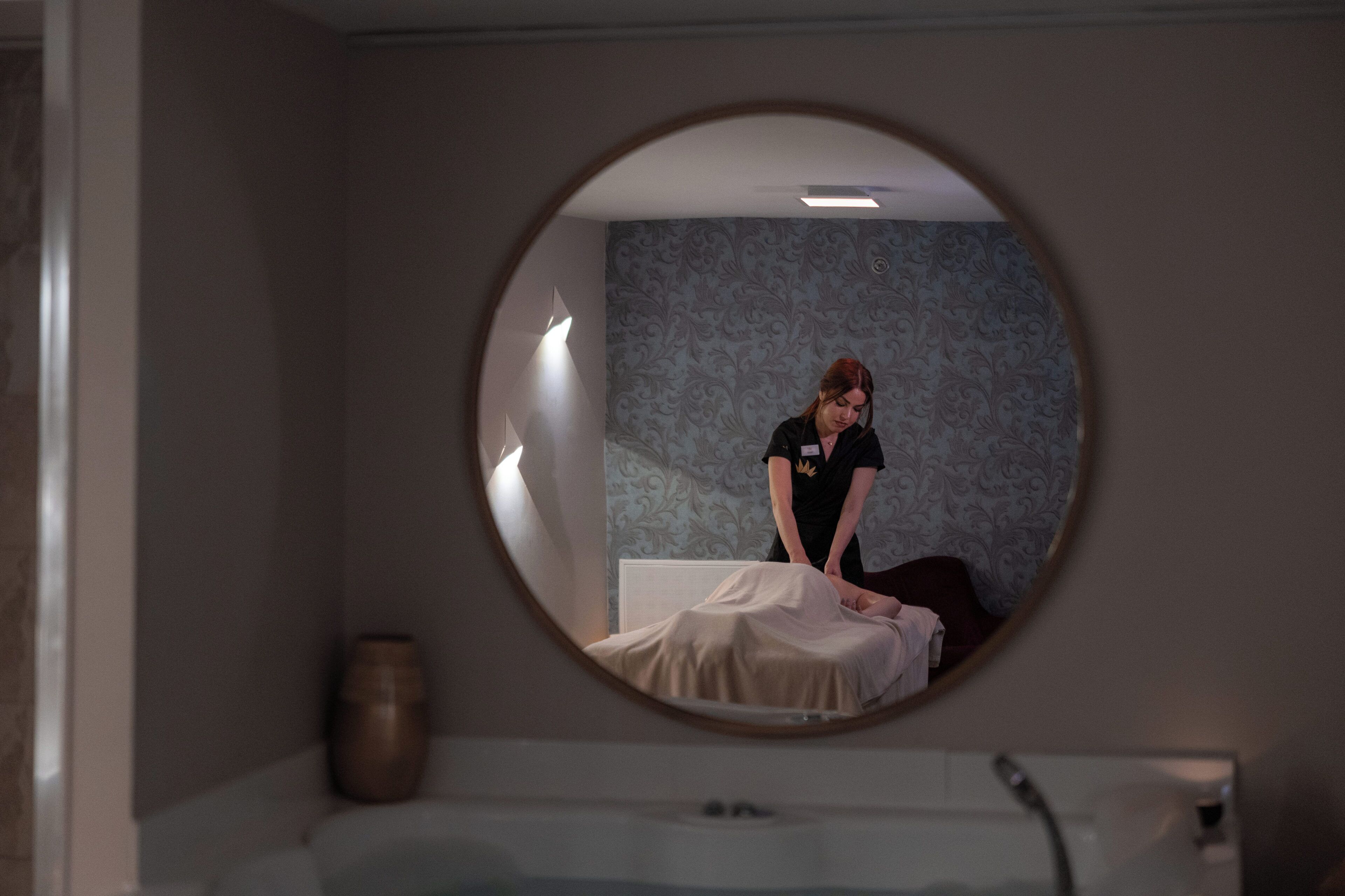 Een kamer met een ronde spiegel, een bed, een massagetafel en een wand met behangpapier met een patroon.
