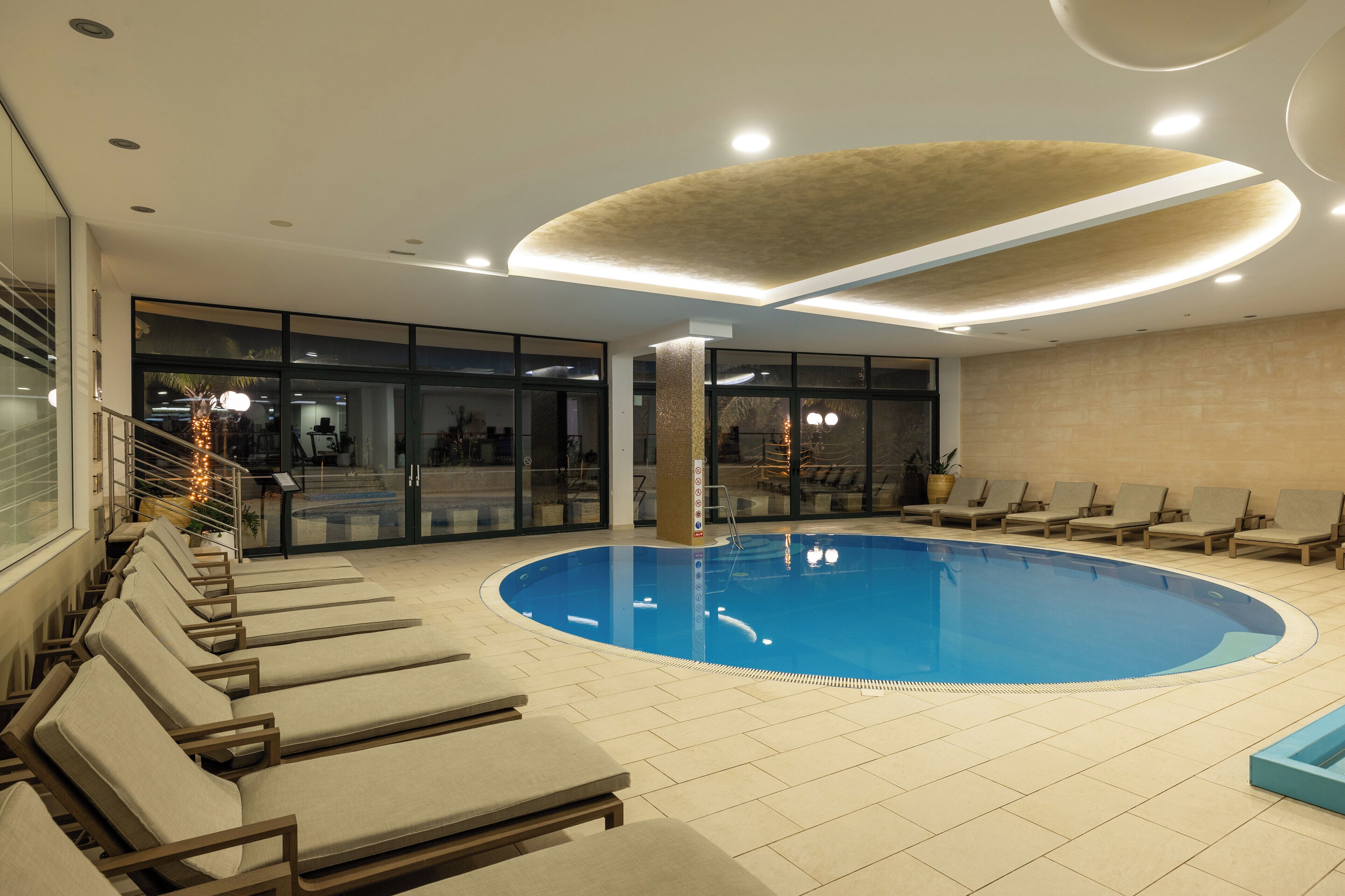 En indendørs swimmingpool med liggestole og en glasvæg.