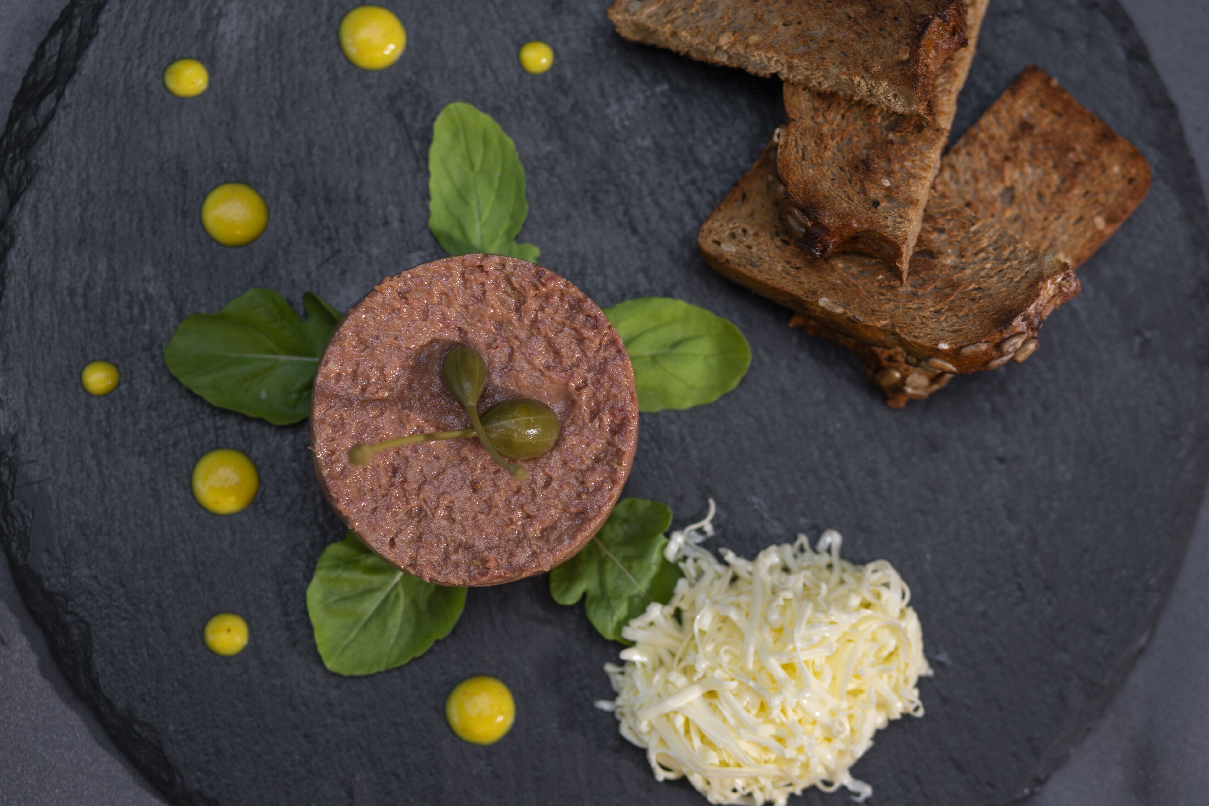 A dark slate plate with a round meat slice, green leaves, yellow dots, shredded cheese, and slices of bread.