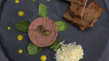 A dark slate plate with a round meat slice, green leaves, yellow dots, shredded cheese, and slices of bread.