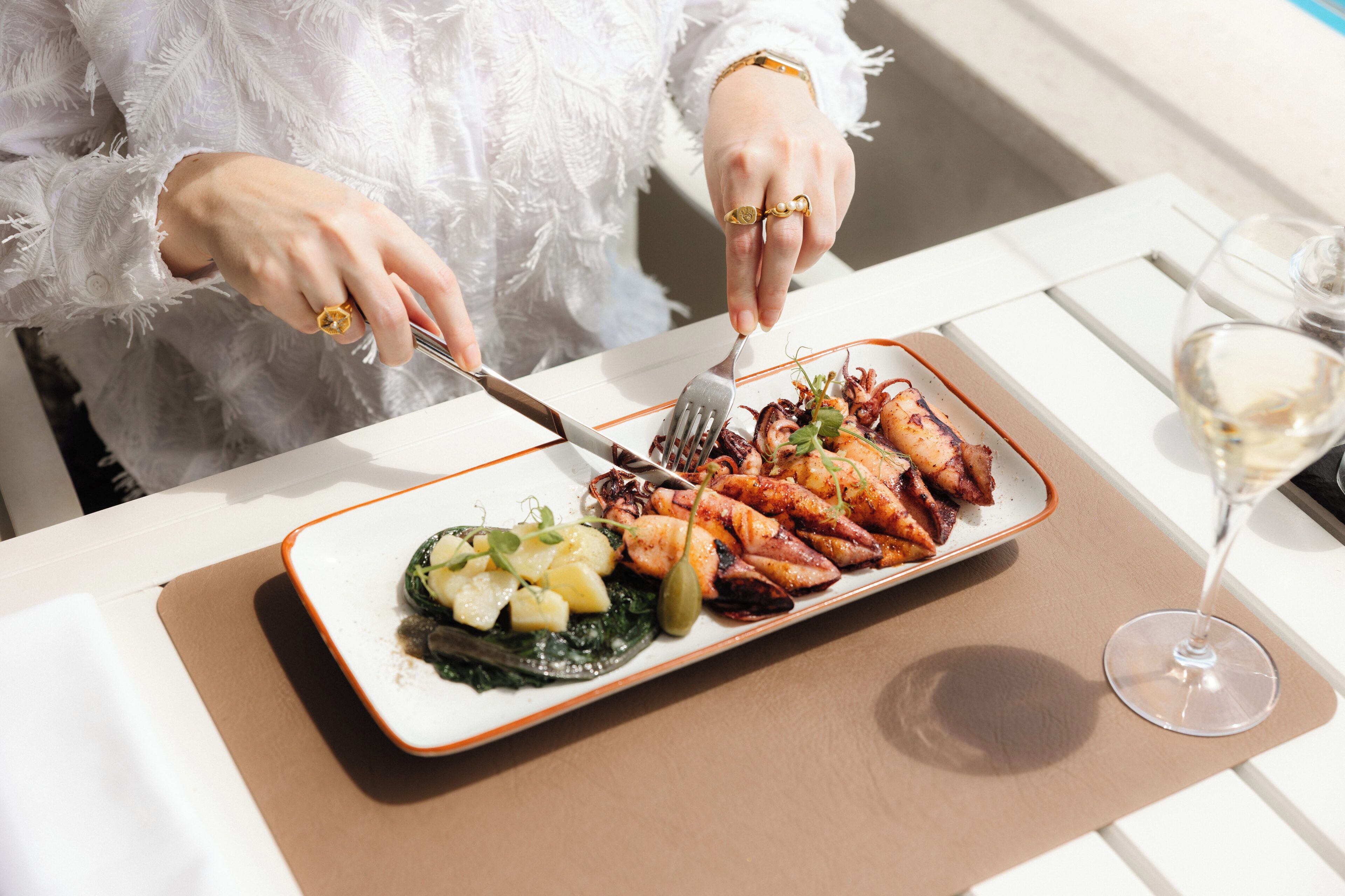 A person dining at a table with a plate of food, a fork, and a glass of wine.
