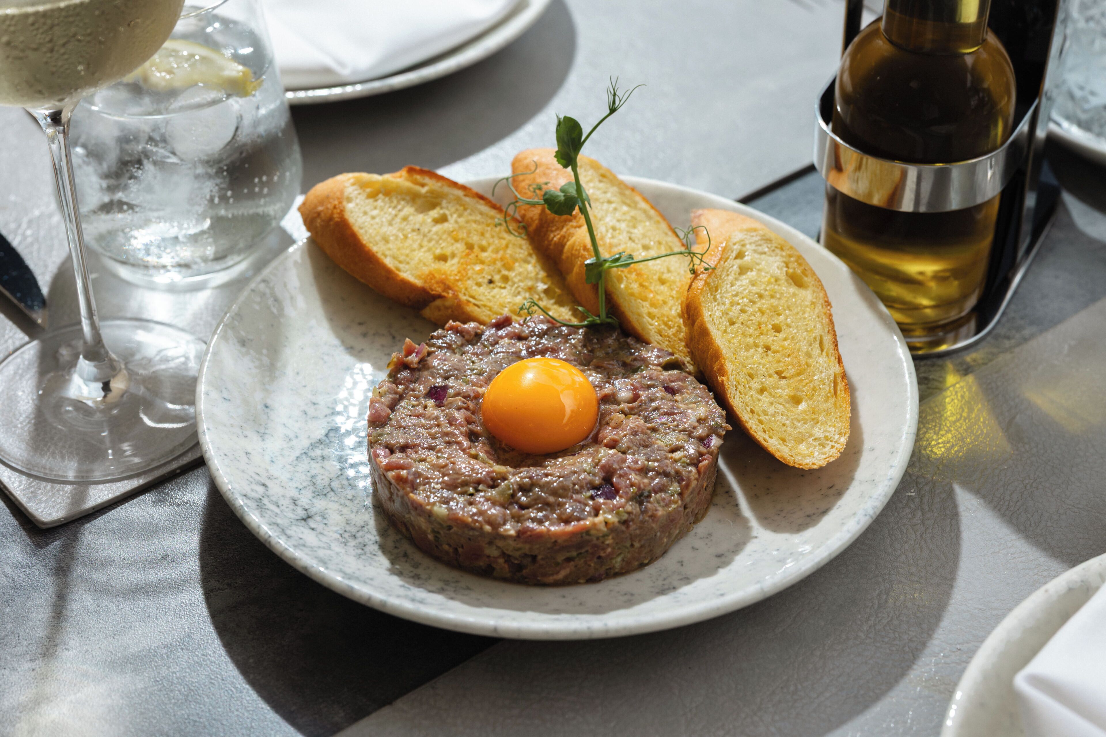 A plate with a meat-based dish, a glass of water, and slices of bread on a table.