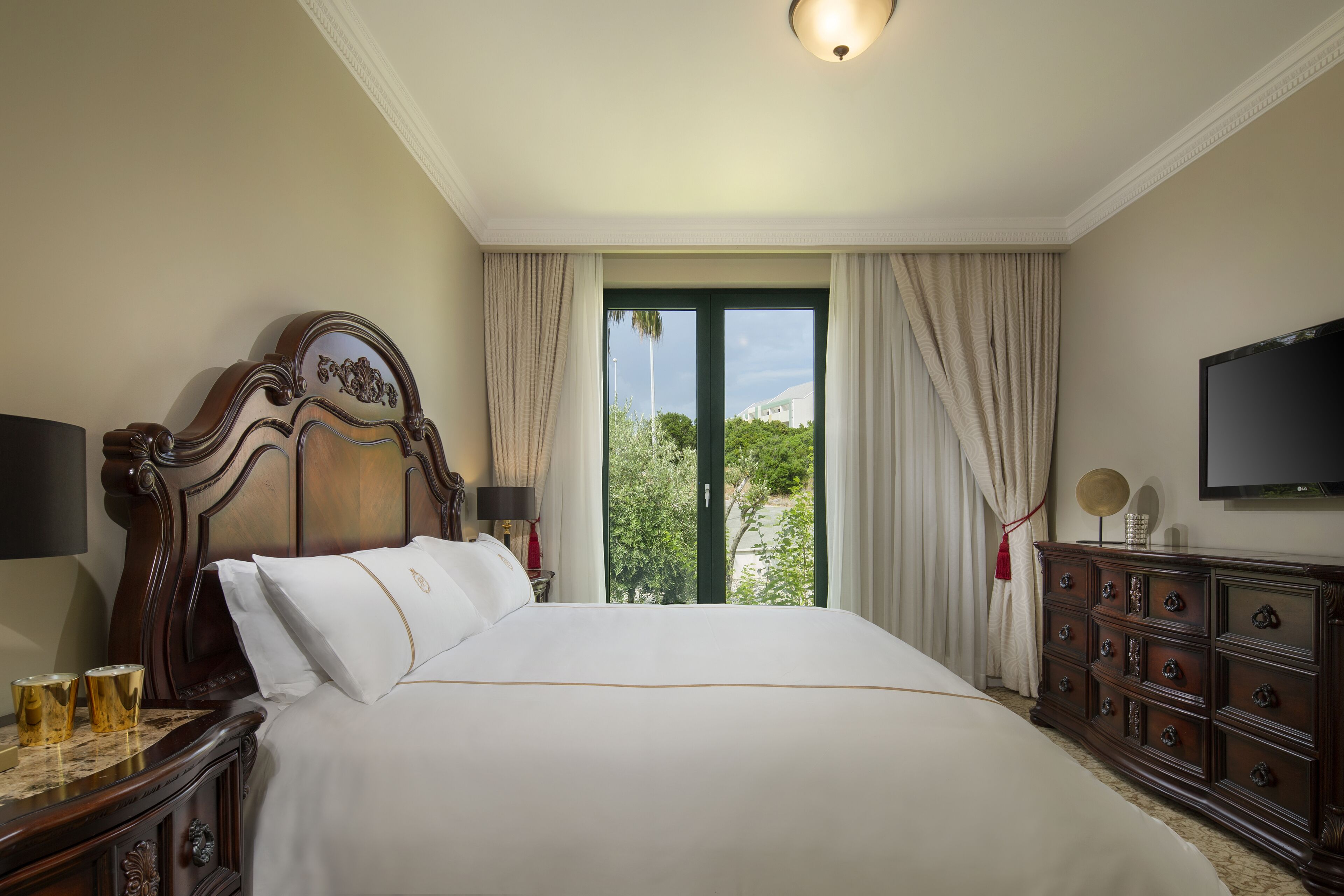 A hotel room with a large bed, wooden headboard, a dresser, a television, and a view of greenery outside.
