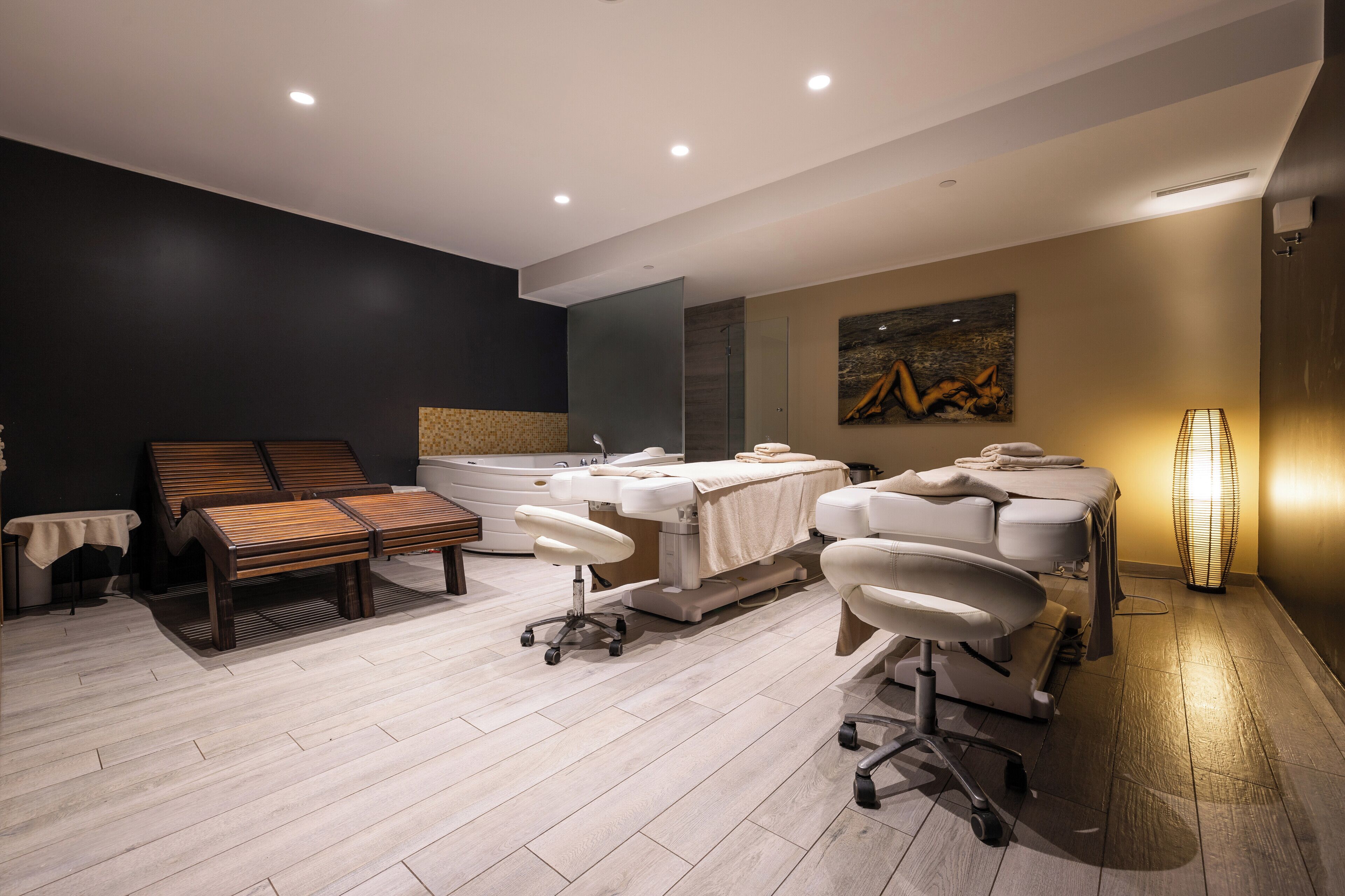 A modern spa room with a massage table, two massage chairs, a large bathtub, and a wall-mounted massage table.