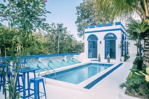 A pool area with a white and blue building, a clear blue pool, and surrounding greenery. - Bombora Surf Camp (Pecatu)