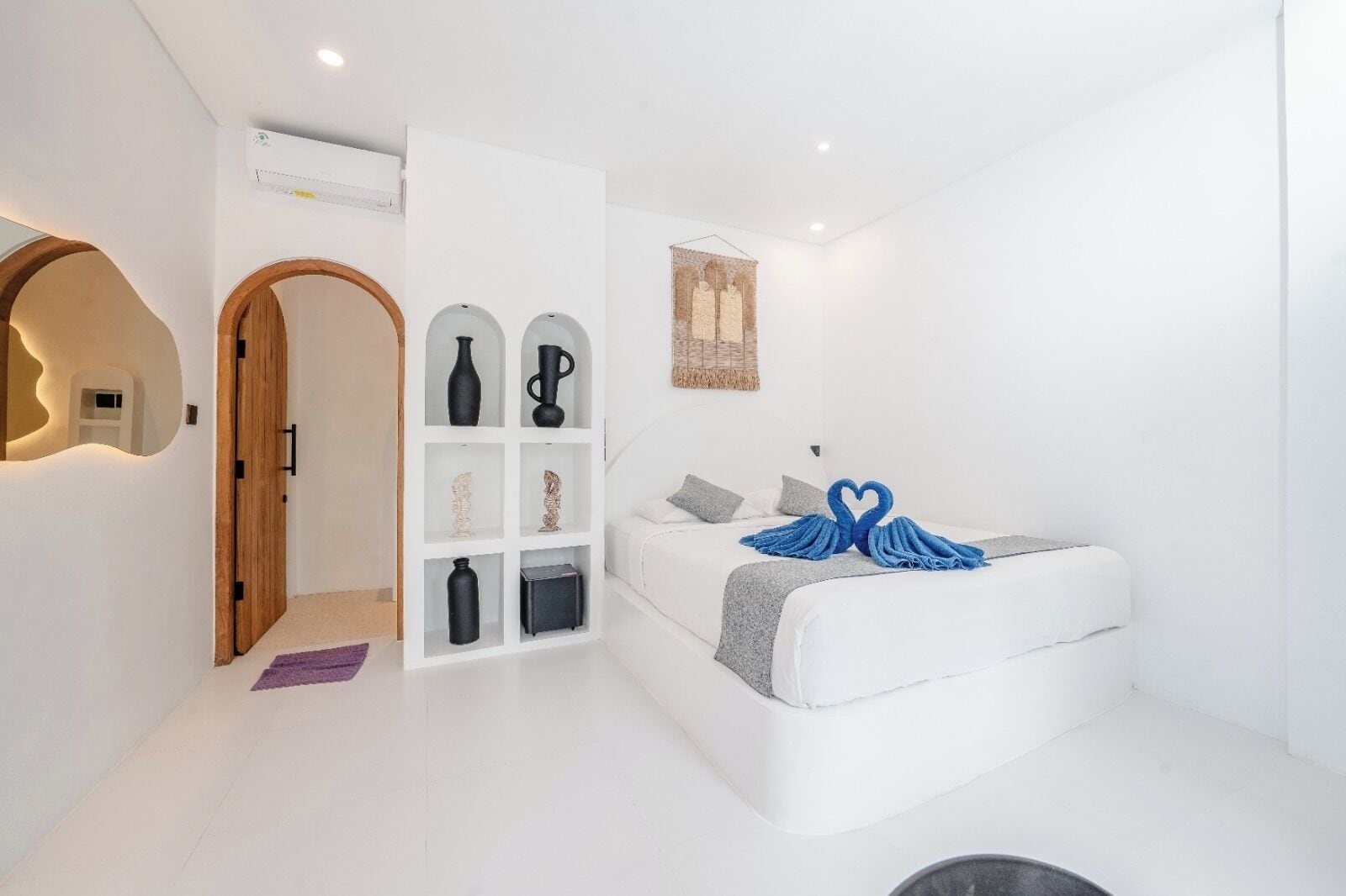 A modern, white-themed bedroom with a built-in wardrobe, a bed with a heart-shaped towel, and a wall-mounted air conditioning unit.