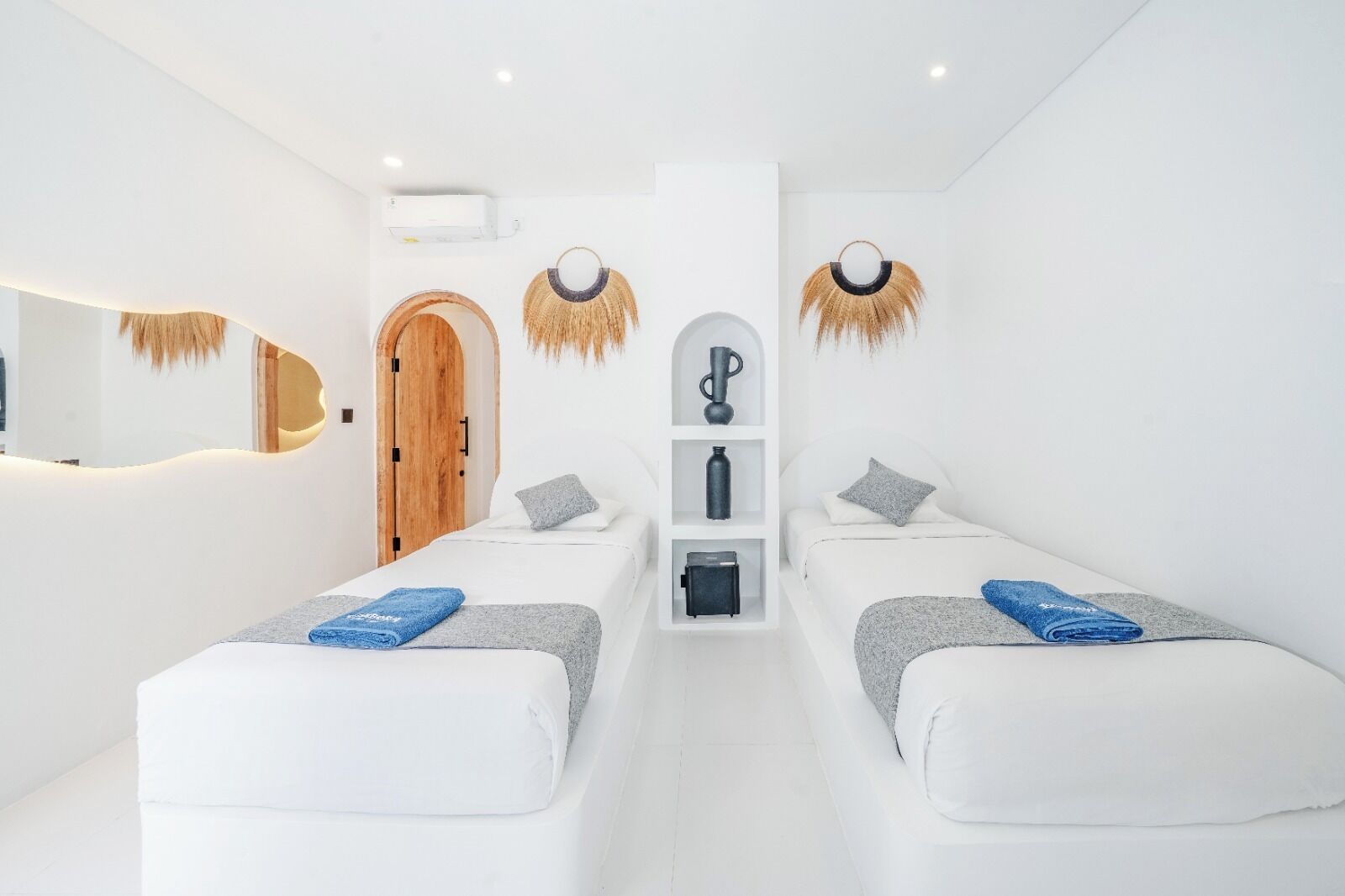 Una habitación de hotel moderna con dos camas, una estantería central con objetos decorativos, una puerta de madera y una pared blanca.