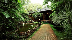 A pathway leading to a traditional wooden structure with a tiled roof, surrounded by lush greenery and a small pond. - The Tree House Resort (Amer)