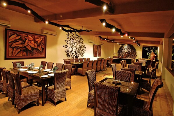 A restaurant with a central dining area featuring round tables and wicker chairs, surrounded by walls adorned with artwork and a large tree sculpture. - The Tree House Resort (Amer)