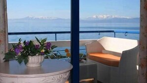 Un balcon avec un canapé en osier blanc, une petite table sur laquelle est posé un vase de fleurs, et une vue sur la mer et les montagnes lointaines.