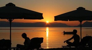 Um pôr do sol sobre um lago tranquilo, com dois guarda-sóis e cadeiras viradas para a água.