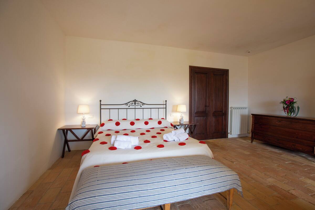 A neatly made bed with rose petals and towels, two bedside tables with lamps, a wooden dresser, and a radiator.