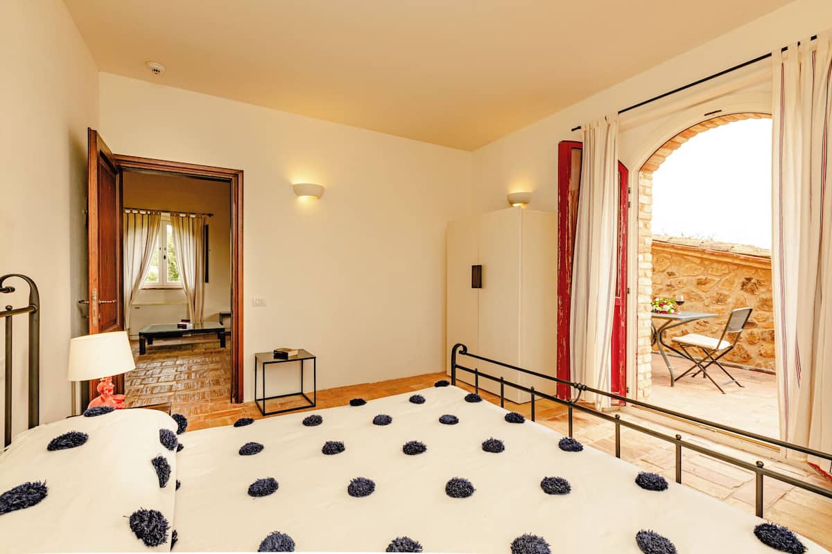A bedroom with a bed, a small table, a chair, a lamp, and a view of an outdoor patio.