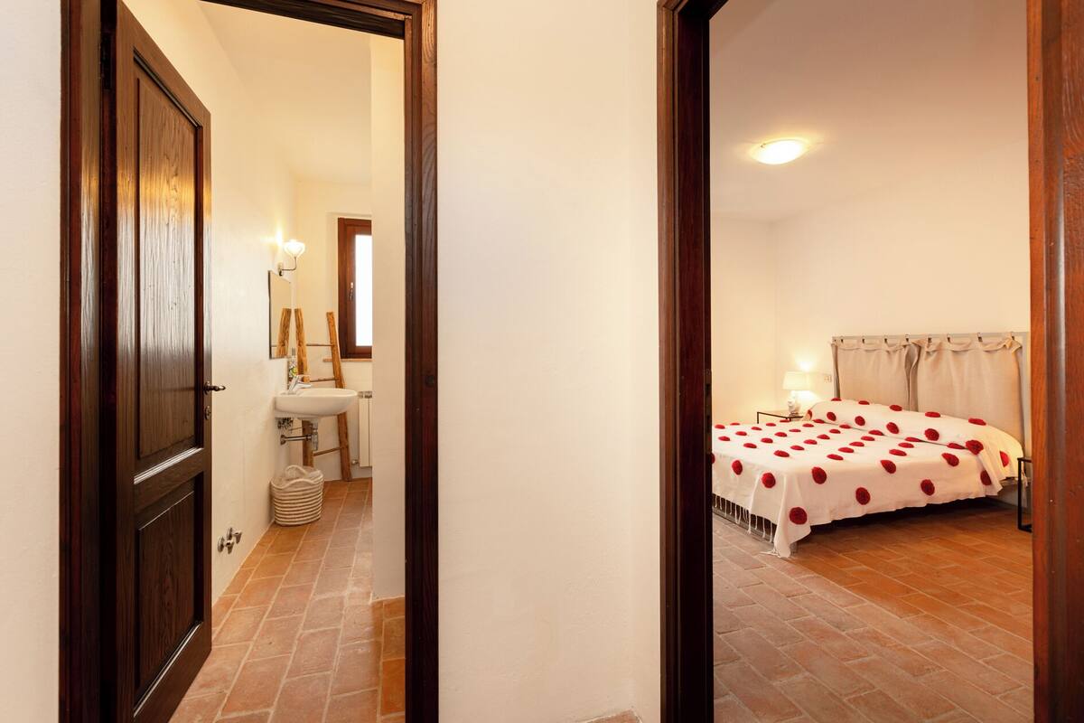 A bedroom with a bed covered in a white sheet with red rose petals, a bedside table with a lamp, and a window with curtains.