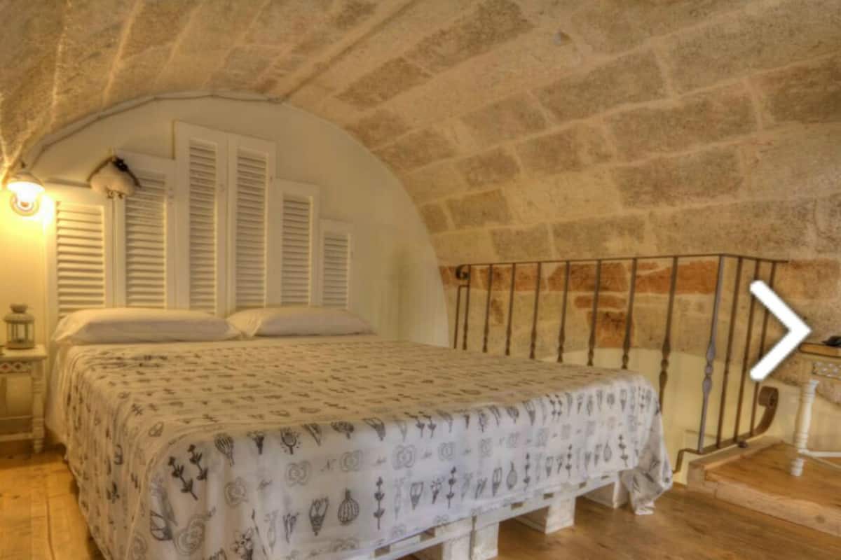 Un dormitorio acogedor con techo inclinado, cama con ropa de cama estampada, mesita de noche con lámpara y ventana con contraventanas.