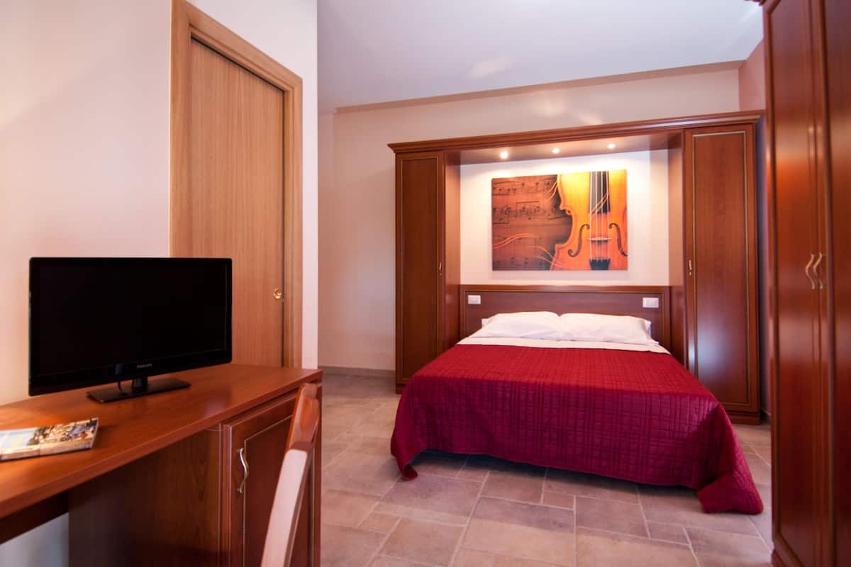 A hotel room with a bed, a television on a wooden desk, and a painting on the wall.