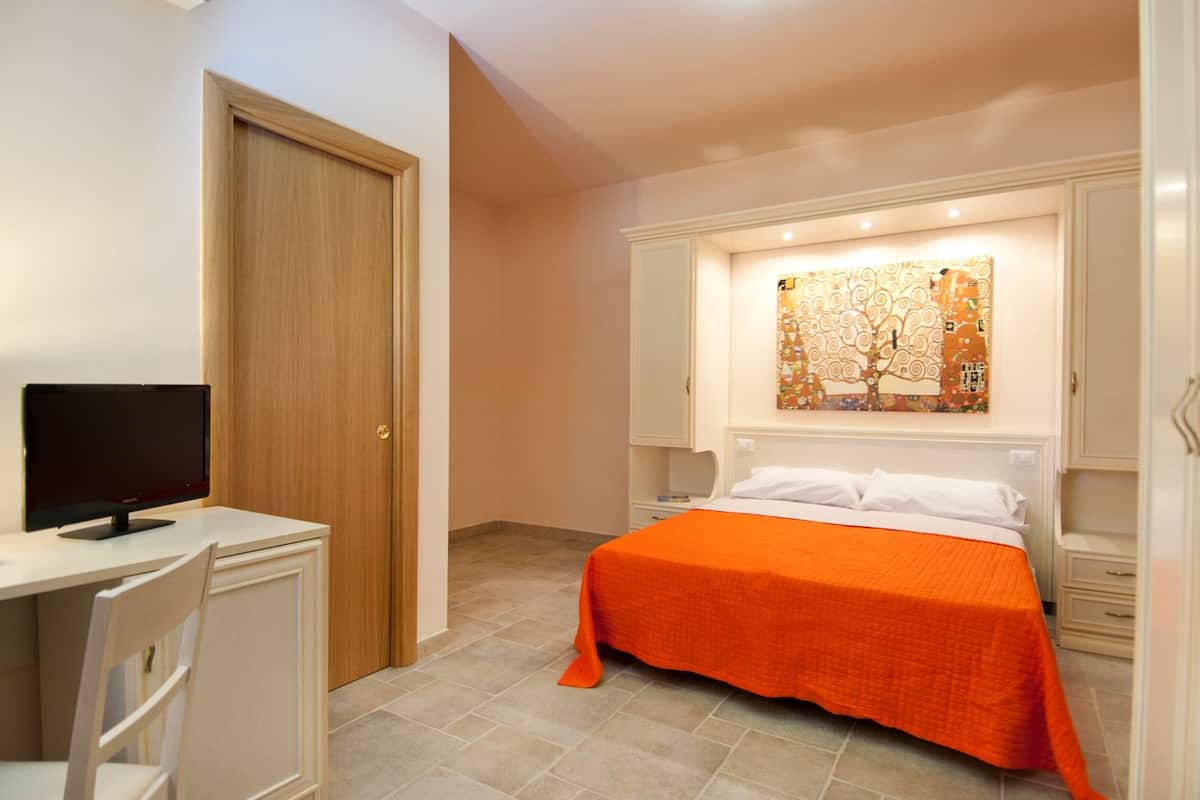 A bedroom with a bed, a desk with a computer, and a painting on the wall.