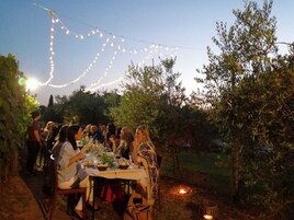 Un área de comedor al aire libre con luces colgantes, mesas preparadas para una comida y un entorno natural.