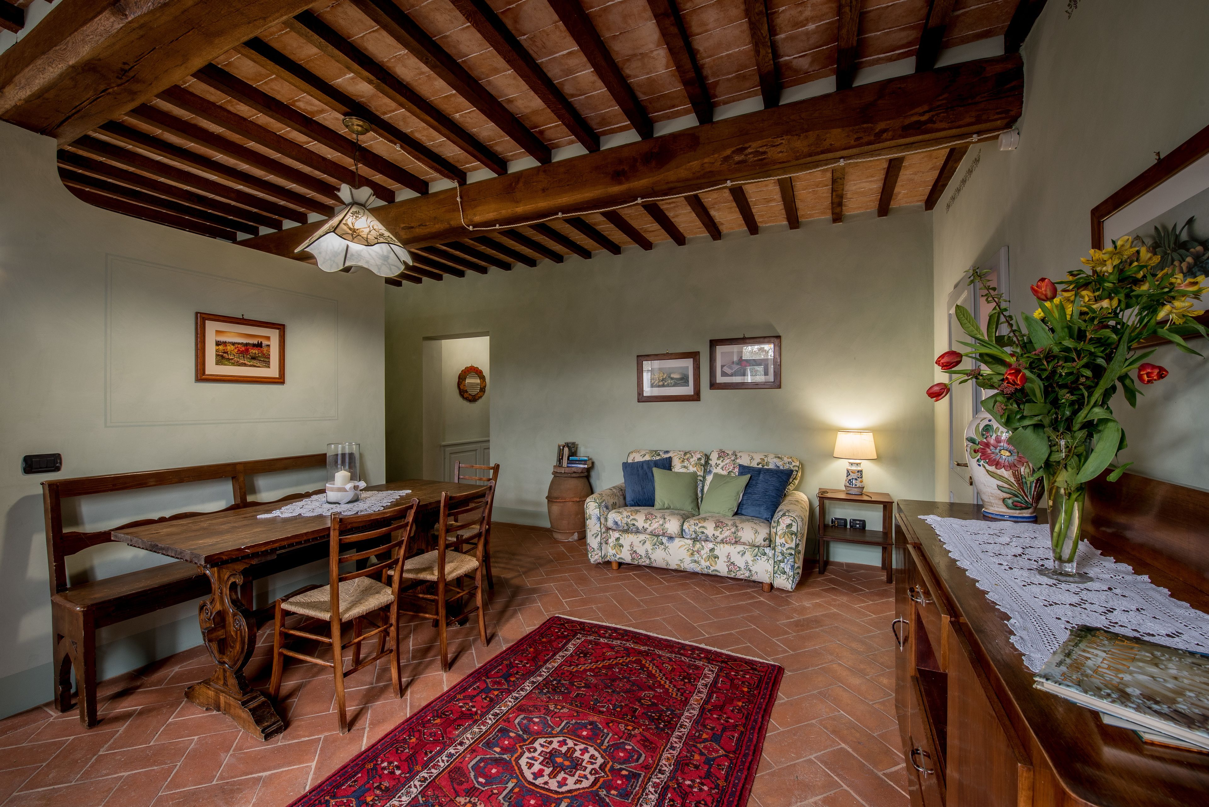 Ein Essbereich mit Holztisch und -stühlen, einer Bank, einem floralen Sofa, einem roten Teppich mit Muster und einer Holzkommode mit einer Blumenvase.