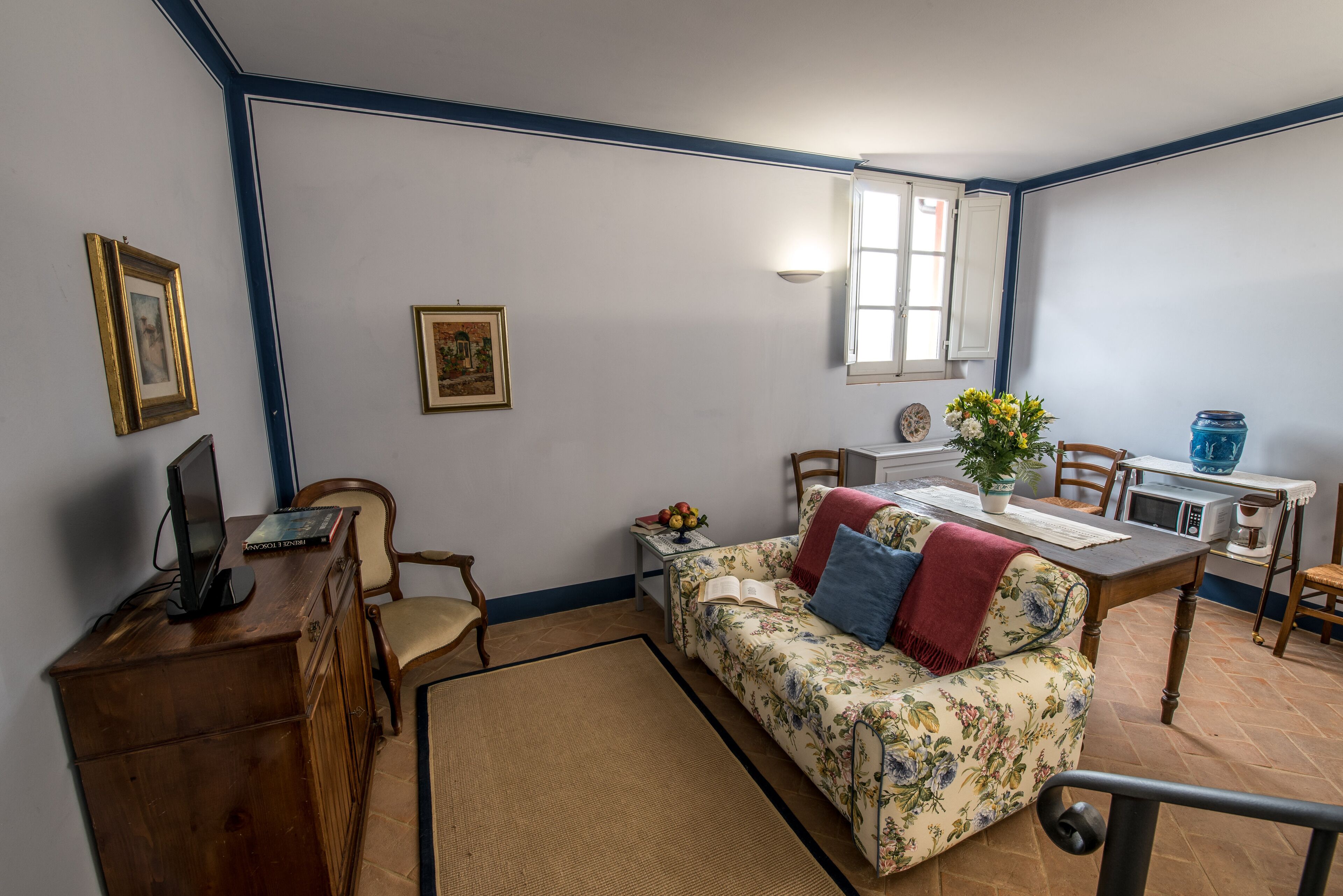 Una sala de estar acogedora con un sofá floral, muebles de madera, una mesa de comedor y una zona de cocina con un microondas y un tarro azul.