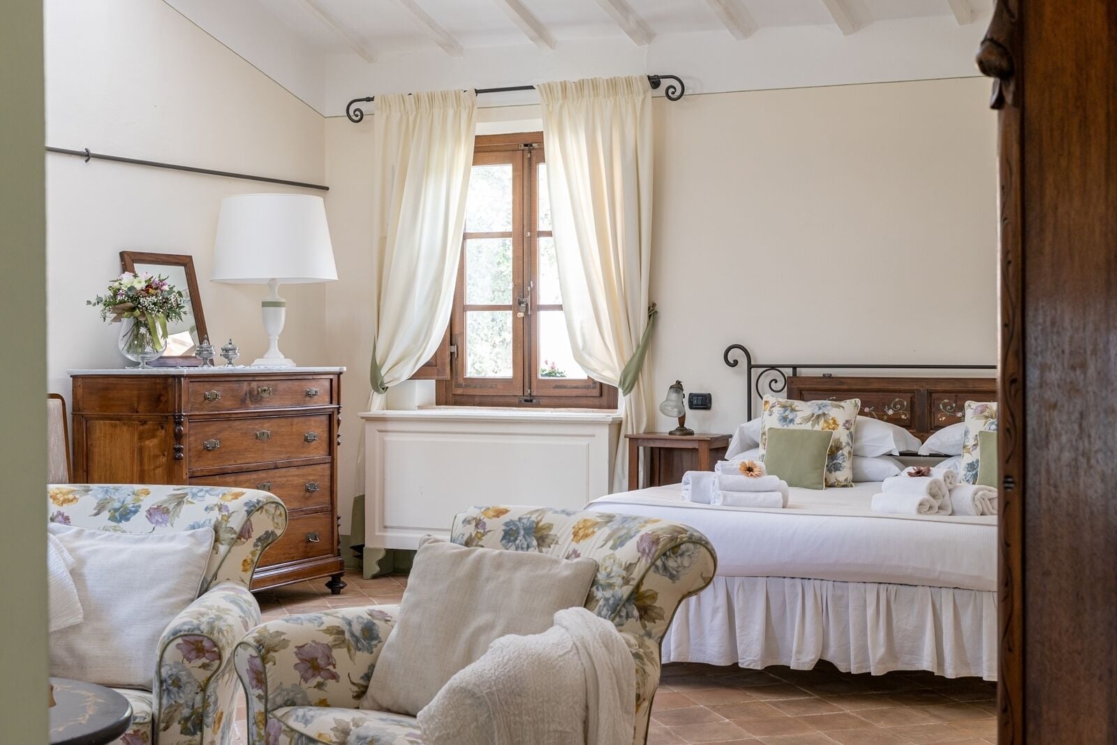 A bedroom with a wooden dresser, a floral armchair, a bed with floral bedding, and a window with curtains.