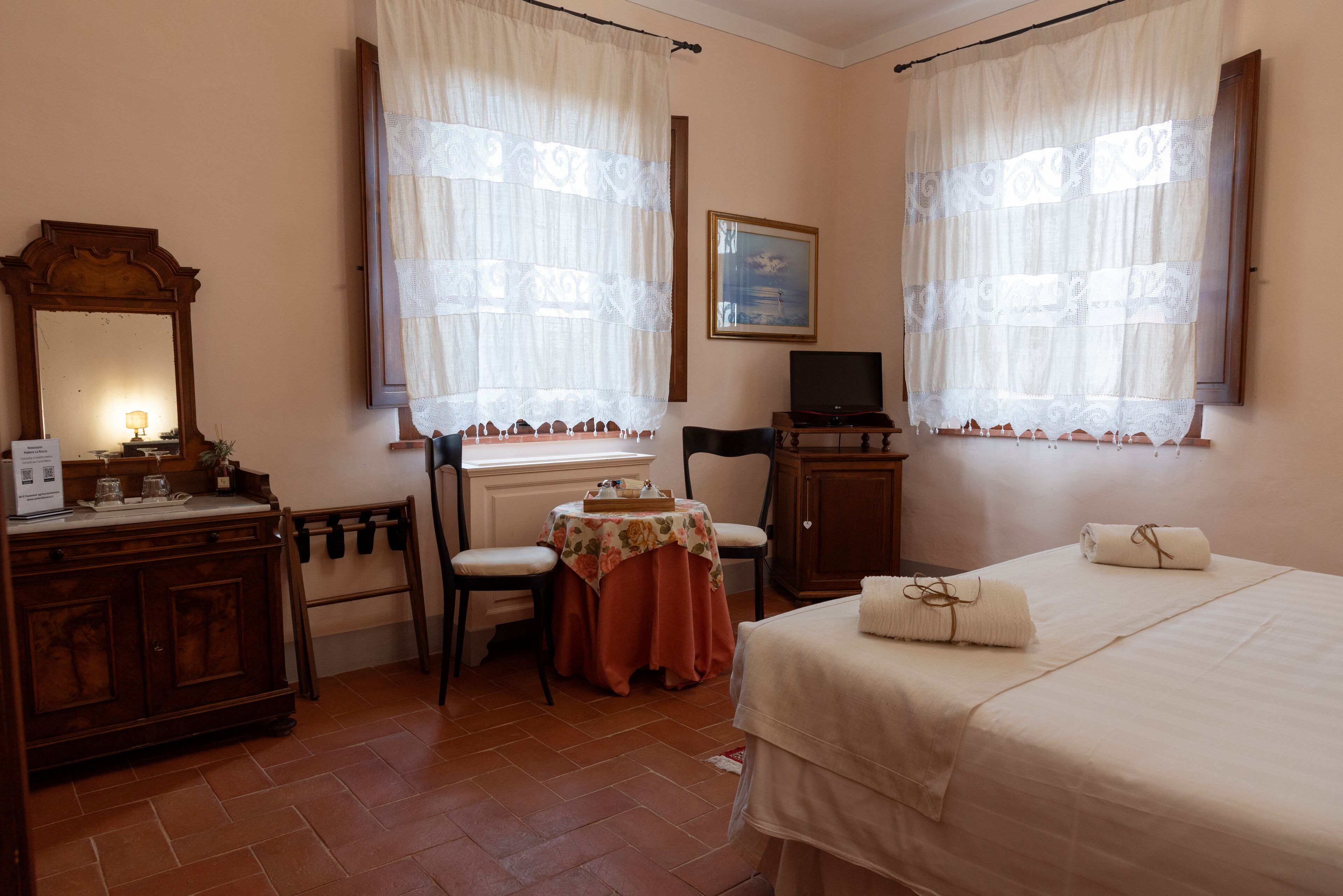 Habitación de hotel con cama, escritorio, silla, mesa con mantel floral, televisión y un cuadro en la pared.