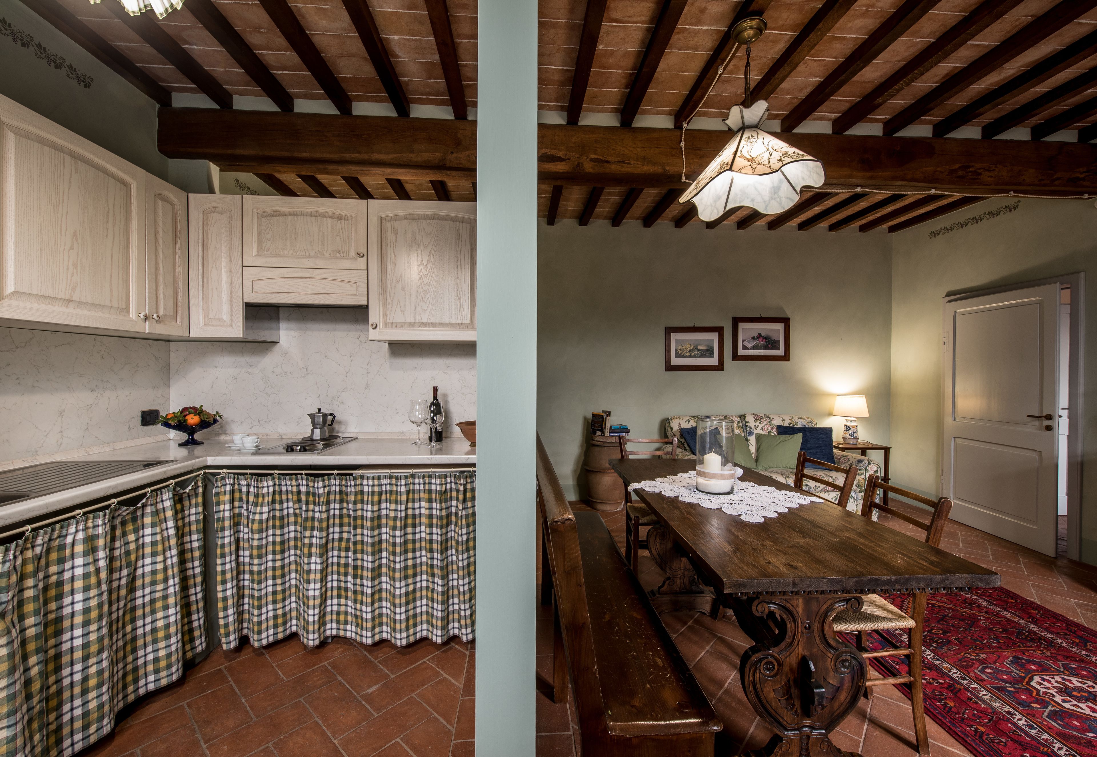 A kitchen with white cabinets and a checkered tablecloth, and a dining area with a wooden table and bench.
