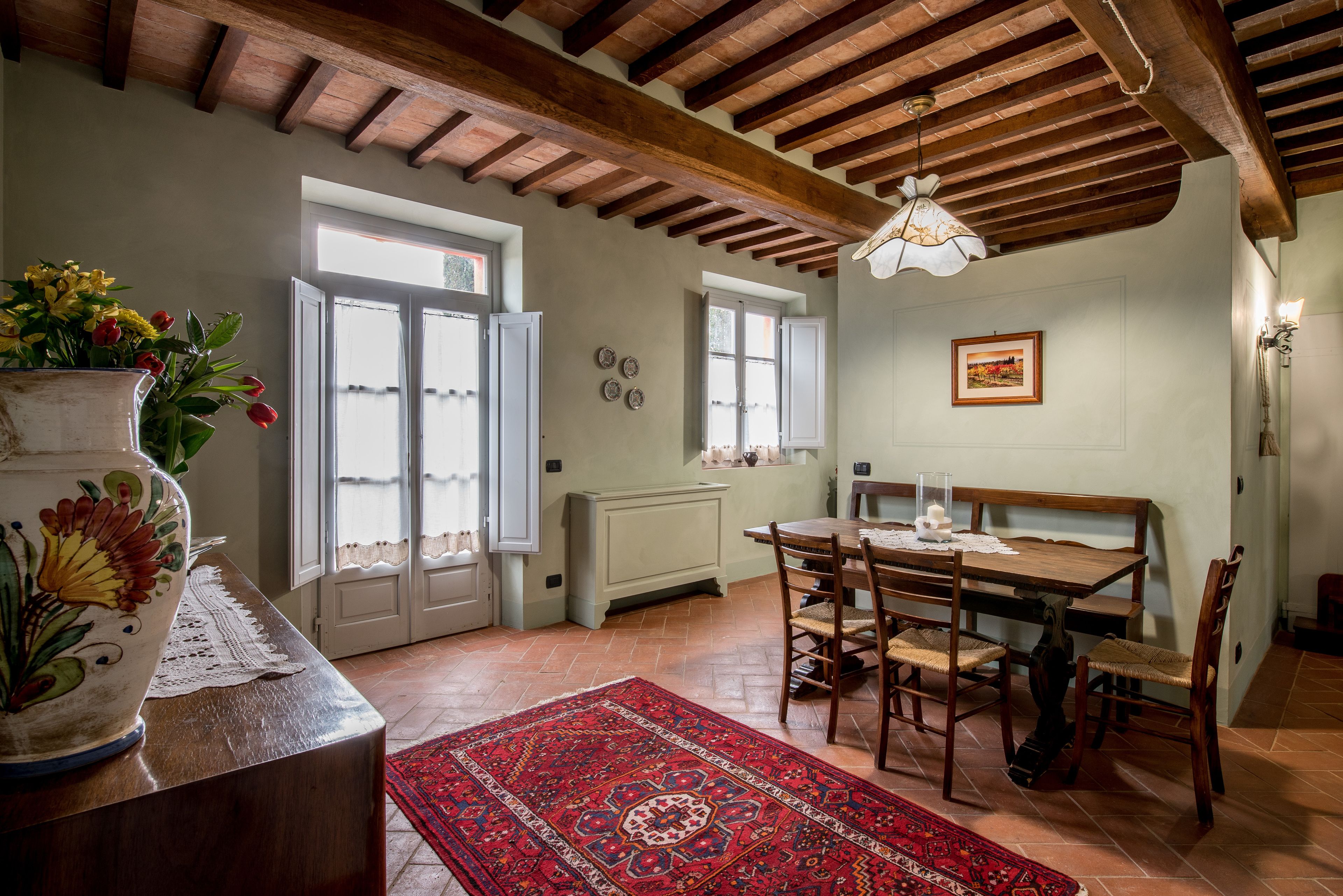 Un'area pranzo con un tavolo e sedie in legno, un tappeto rosso decorato e un vaso bianco con fiori.