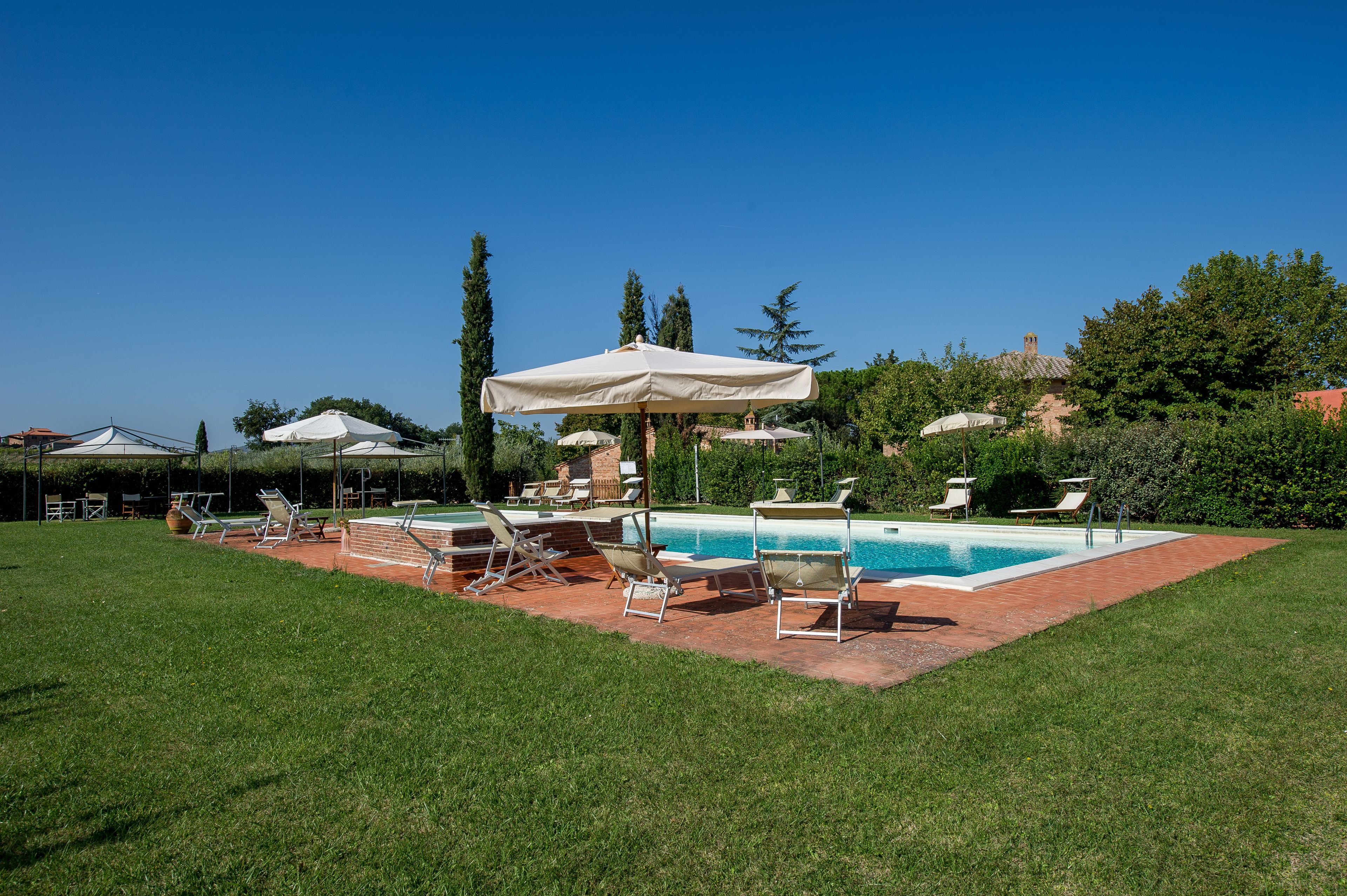 A poolside area with lounge chairs and umbrellas, surrounded by a well-maintained lawn and trees.