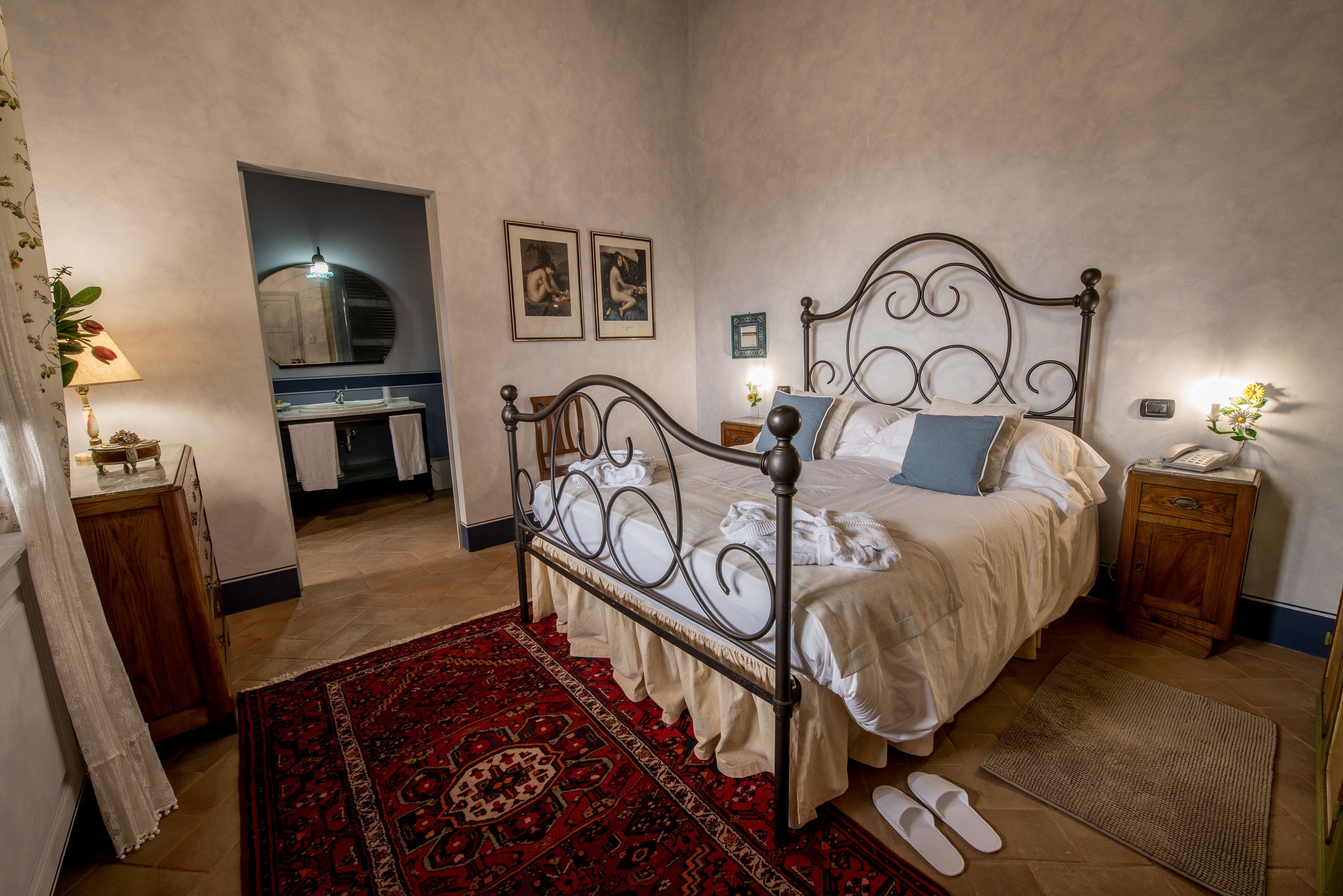 Un dormitorio con estructura de cama metálica, alfombra roja estampada, mesita de noche de madera y una mesita con lámpara.