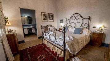 Un dormitorio con estructura de cama metálica, alfombra roja estampada, mesita de noche de madera y una mesita con lámpara.